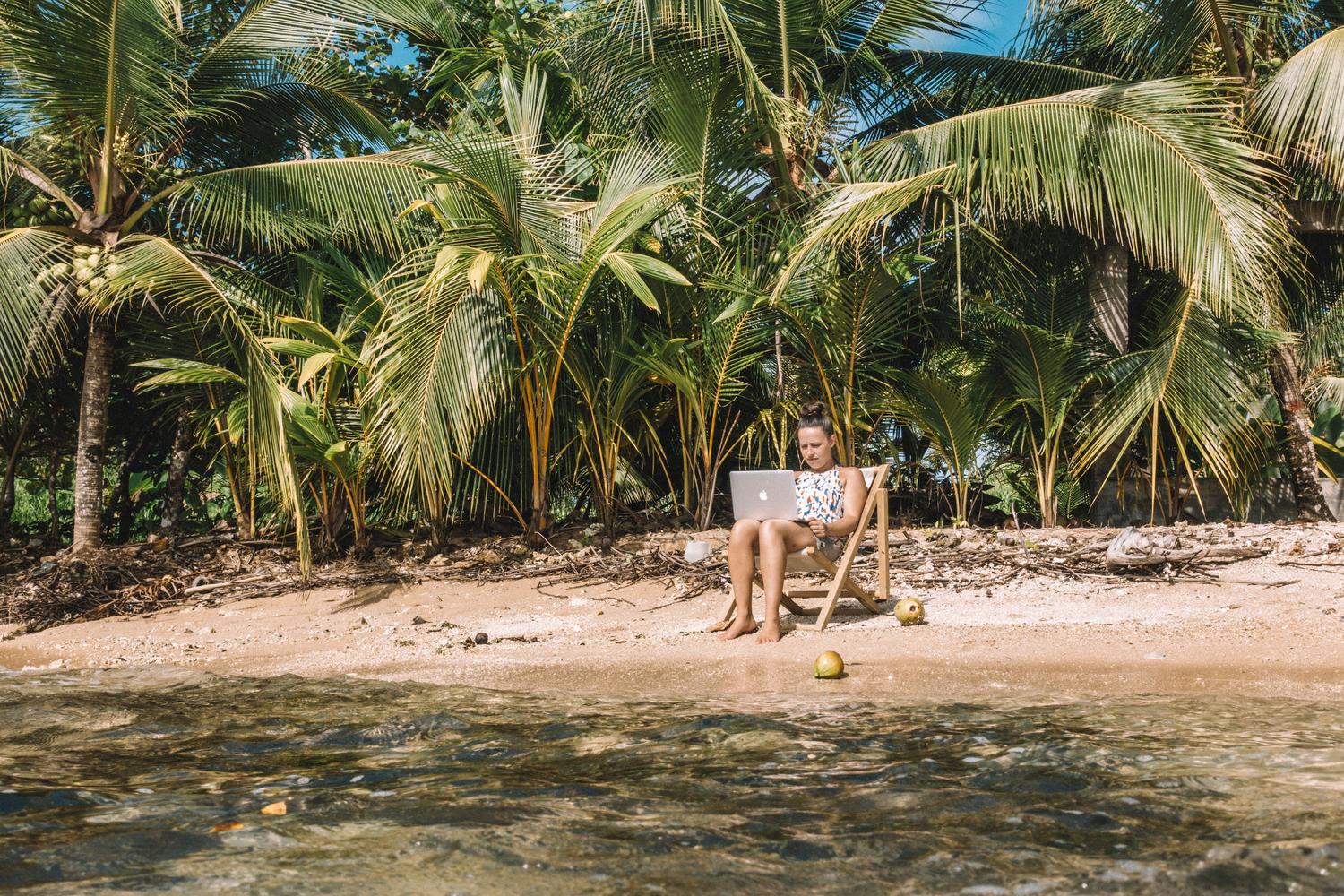 Una mujer trabajando remotamente con un ordenador portátil en una playa tropical, ilustrando el estilo de vida nómada digital.