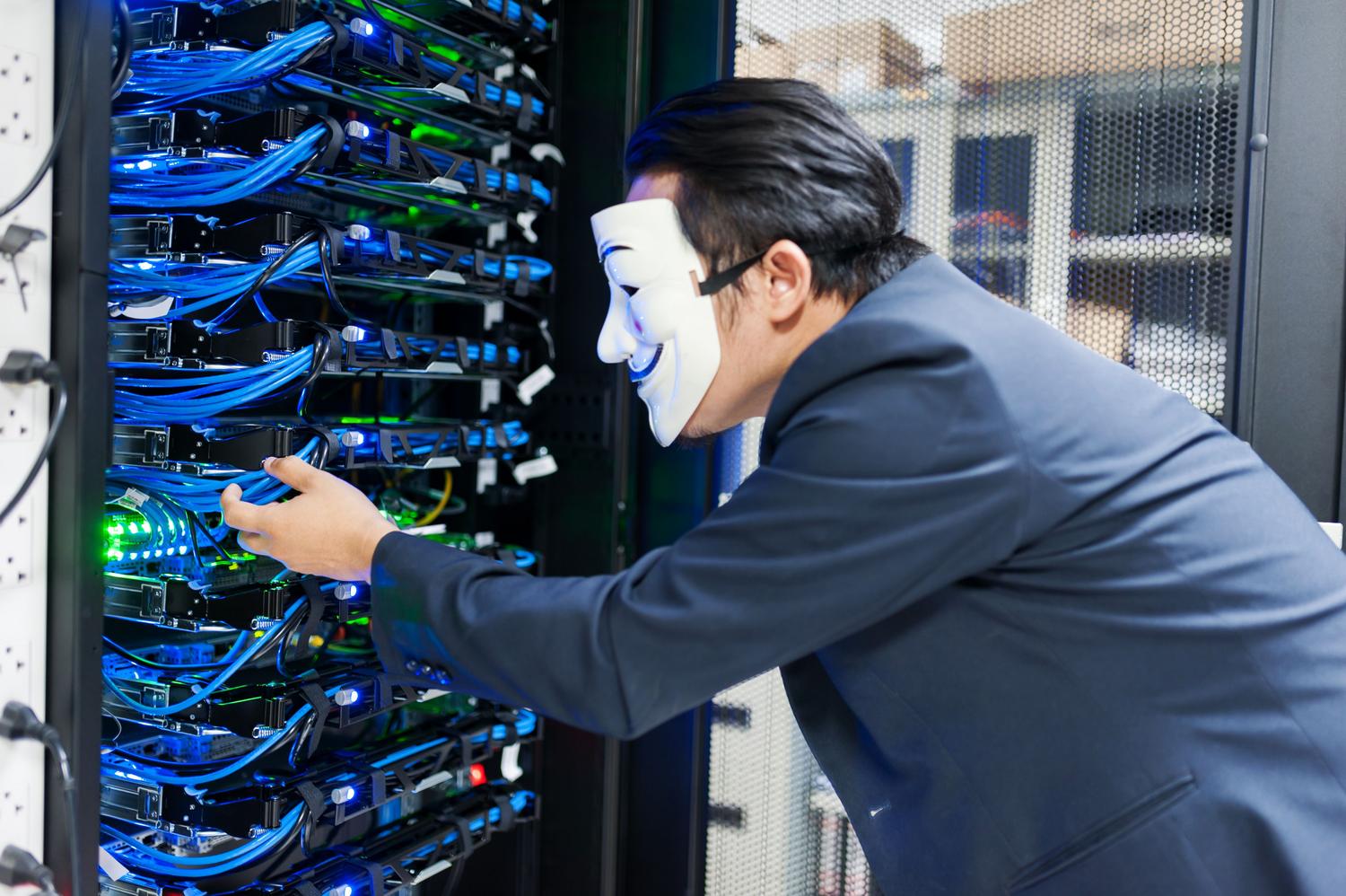 Individuo enmascarado interactuando con racks de servidores en un centro de datos, simbolizando la infraestructura técnica de las Darknets.