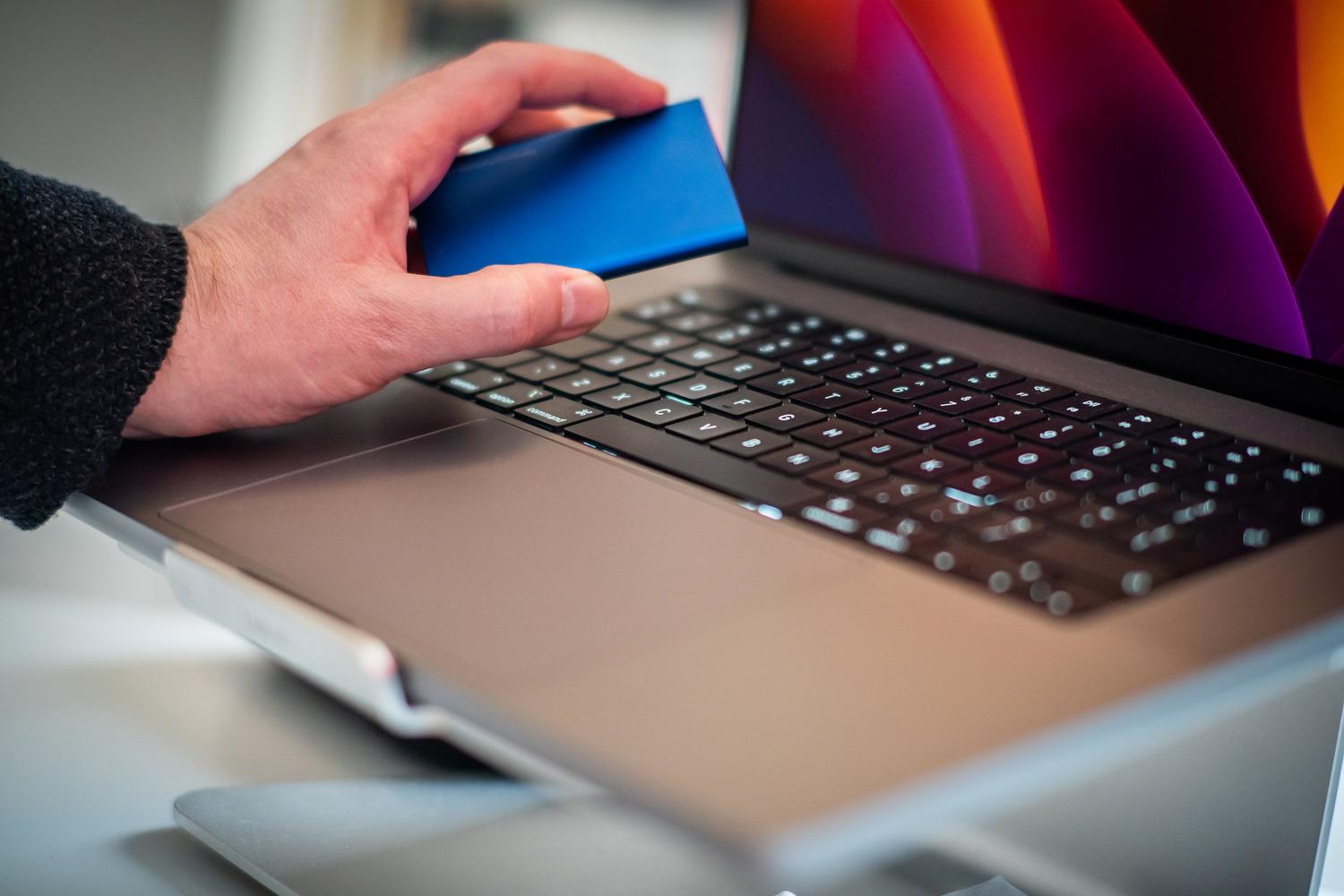 Primer plano de una mano sosteniendo un disco SSD externo junto a un MacBook Pro, representando la transferencia de archivos y el uso de Time Machine.