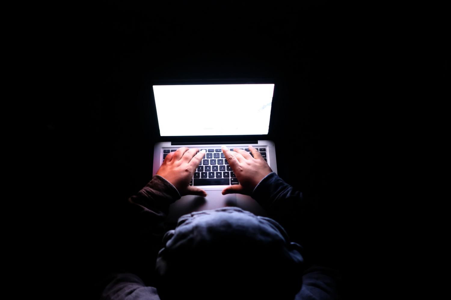 Persona anónima con capucha utilizando un portátil en la oscuridad, representando el peligro invisible de los hackers en redes públicas.