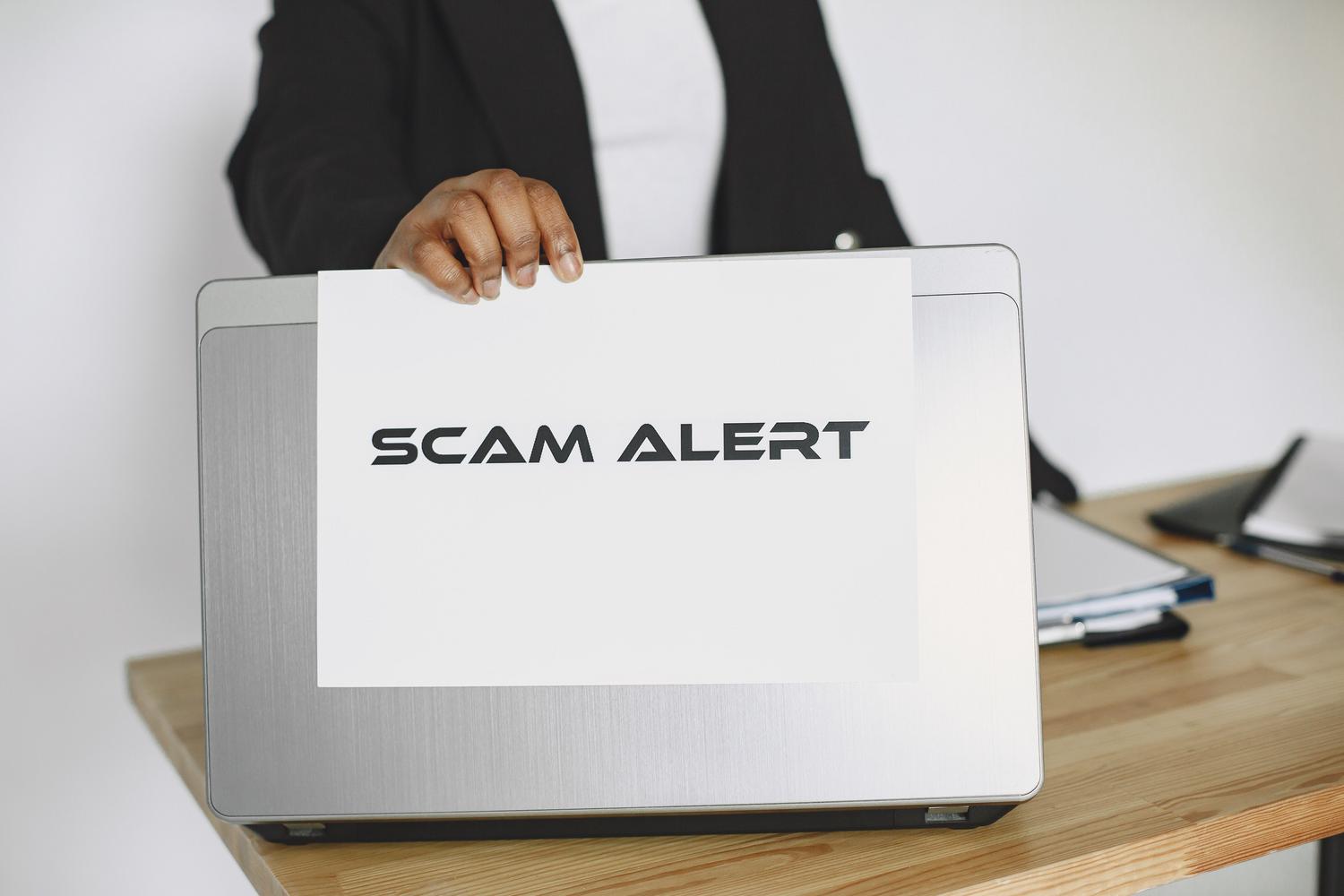 Persona sosteniendo un cartel de 'SCAM ALERT' frente a un portátil, representando las alertas fraudulentas de malware en Chrome.
