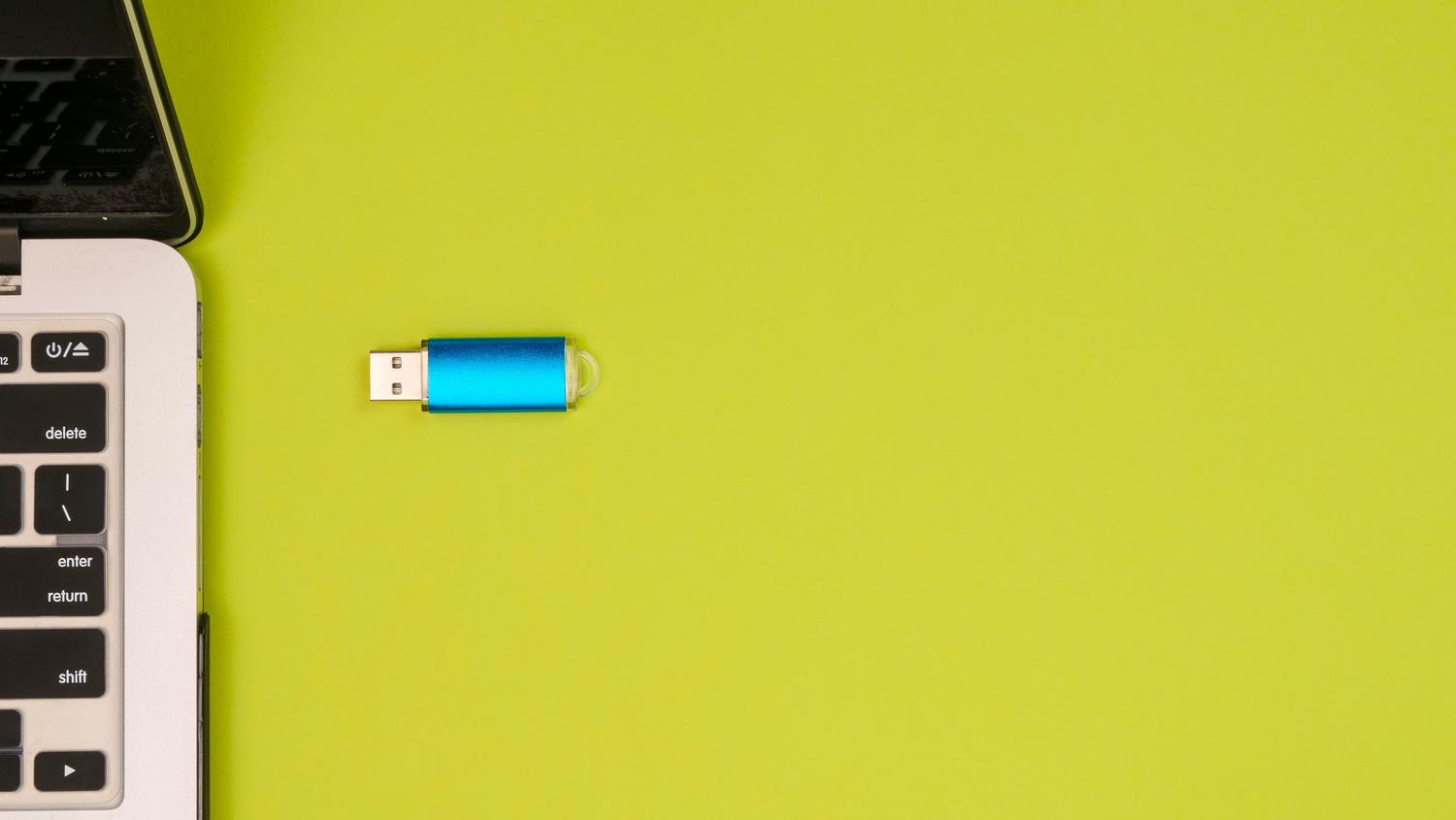 Pendrive USB azul metálico junto a un portátil sobre un fondo verde vibrante, ideal como imagen de cabecera.