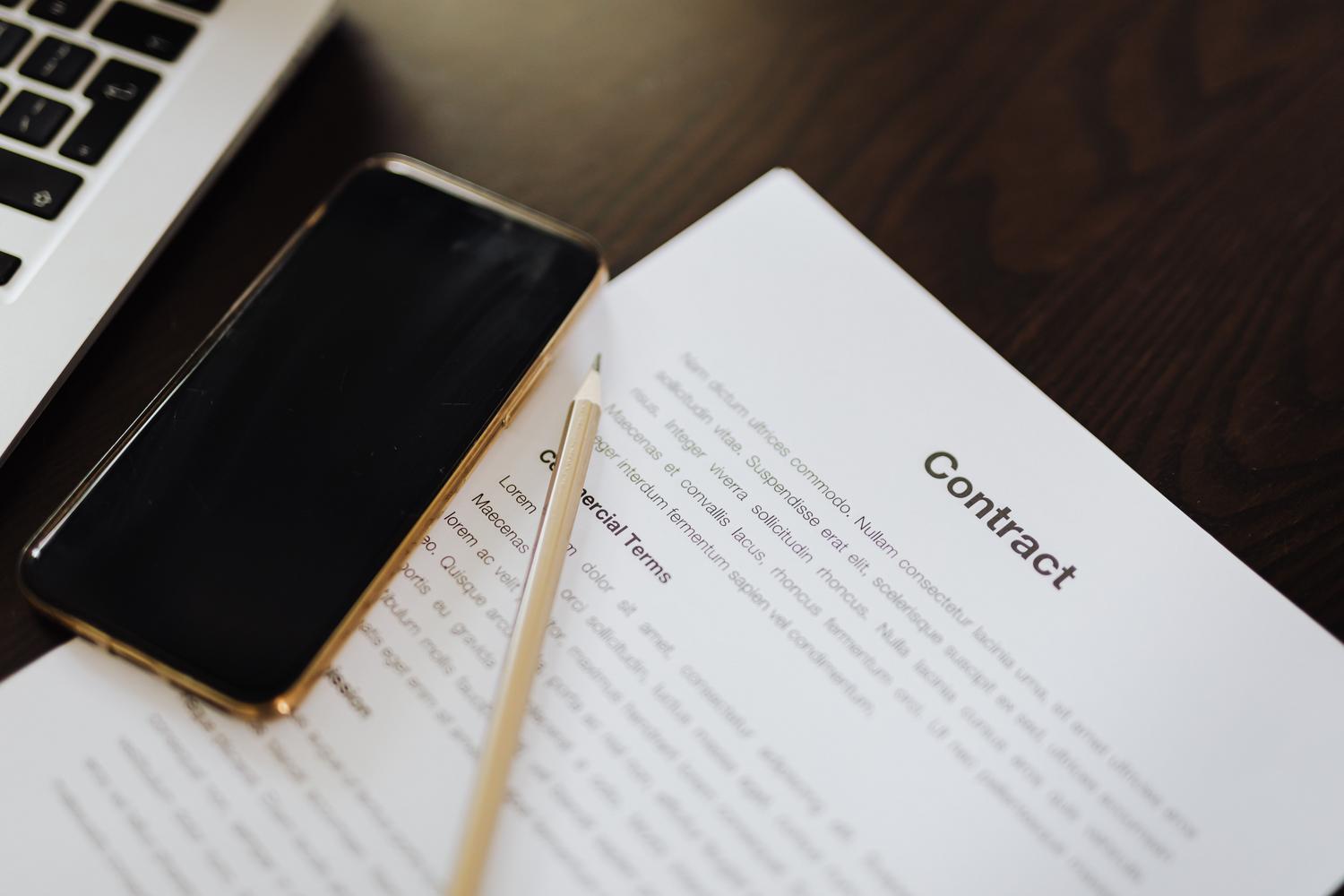 Vista cenital de un contrato impreso junto a un ordenador portátil y un smartphone, simbolizando la gestión digital de documentos.