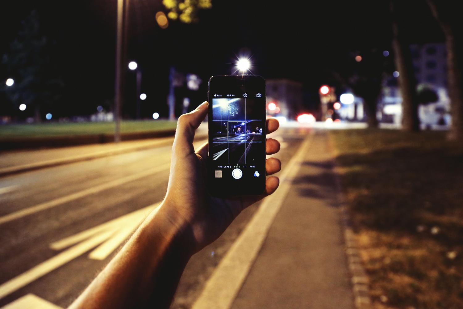 Primer plano de una mano sosteniendo un smartphone para capturar una calle iluminada de noche, ilustrando la fotografía urbana.