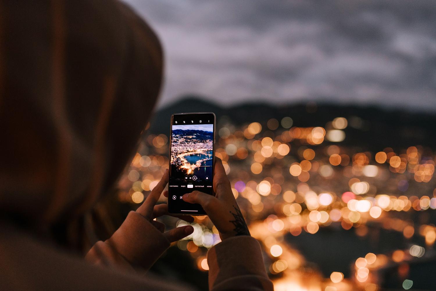 Persona capturando una panorámica nocturna de una ciudad iluminada desde un mirador con su teléfono móvil.