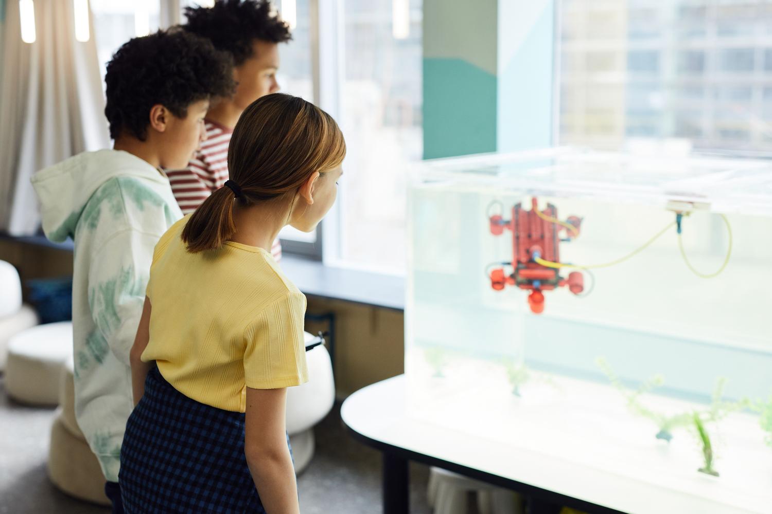 Grupo diverso de niños observando con atención un robot submarino en un tanque de agua, fomentando el trabajo en equipo.