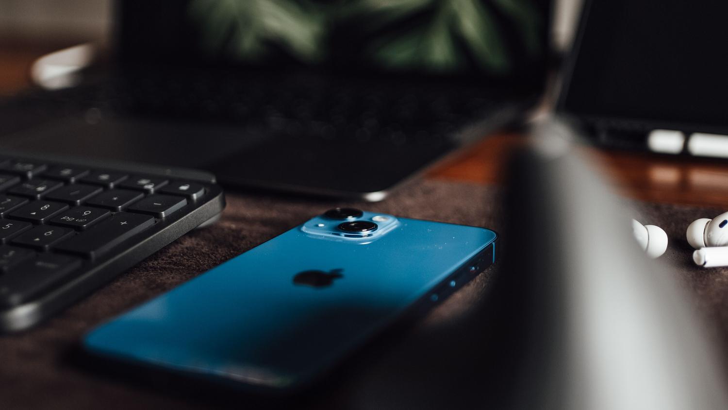 Detalle de un iPhone, teclado de portátil y AirPods sobre una mesa, ilustrando el ecosistema de Apple.
