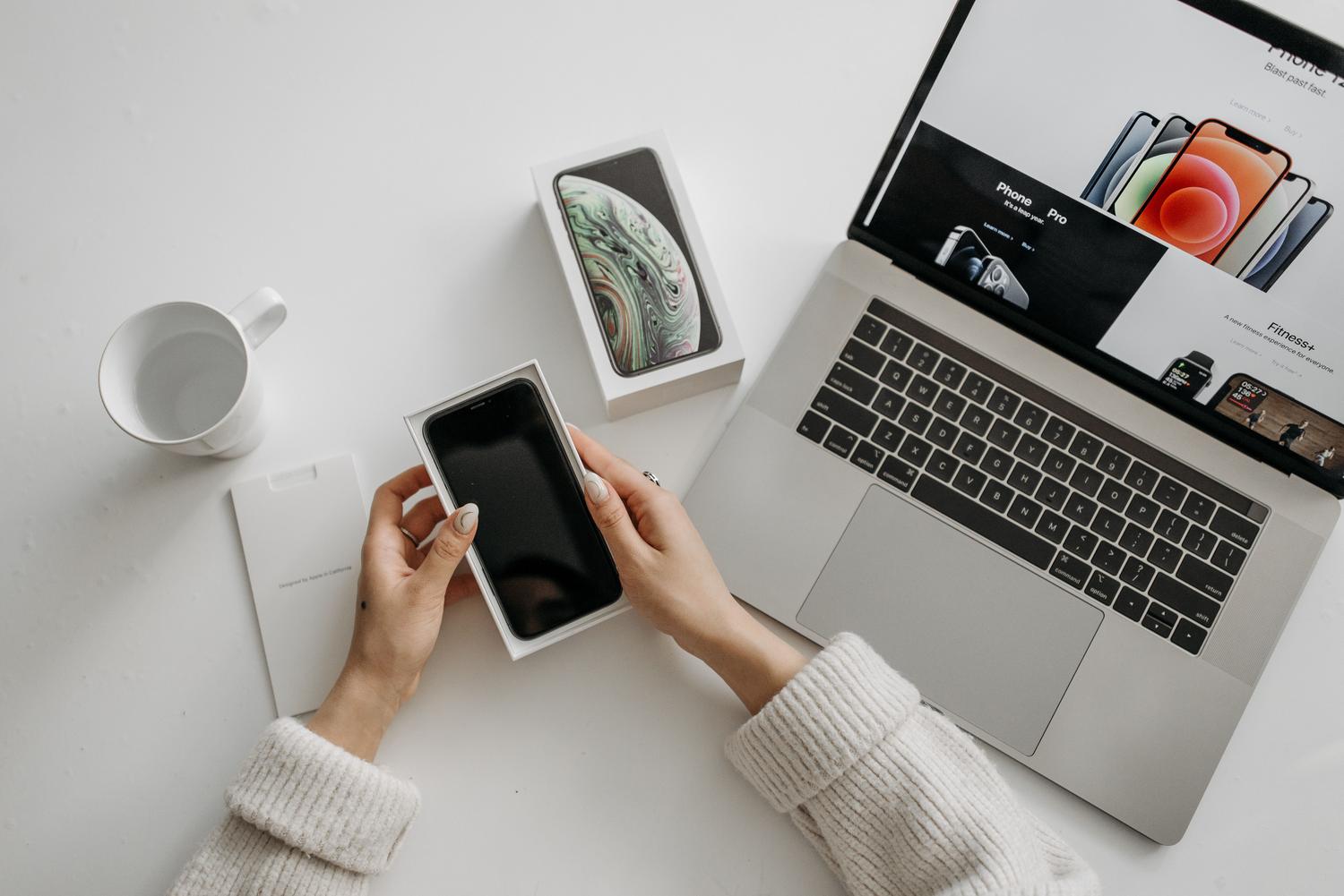 Persona sosteniendo un smartphone junto a un ordenador portátil en un escritorio blanco, mostrando la interacción entre dispositivos.