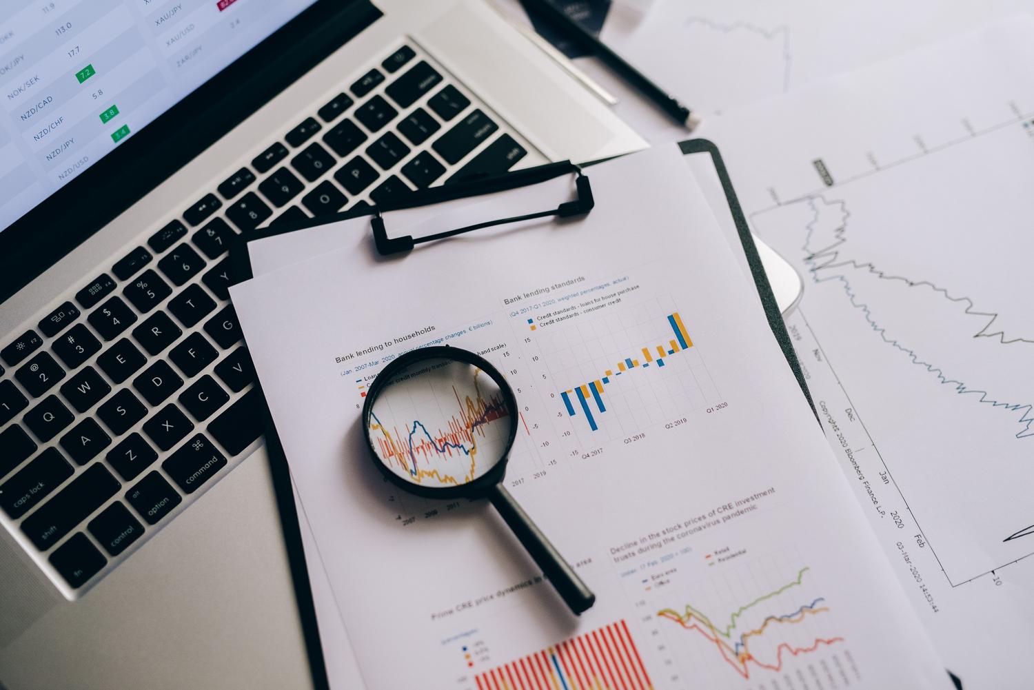 Lupa analizando gráficos de datos financieros impresos junto a un ordenador portátil.